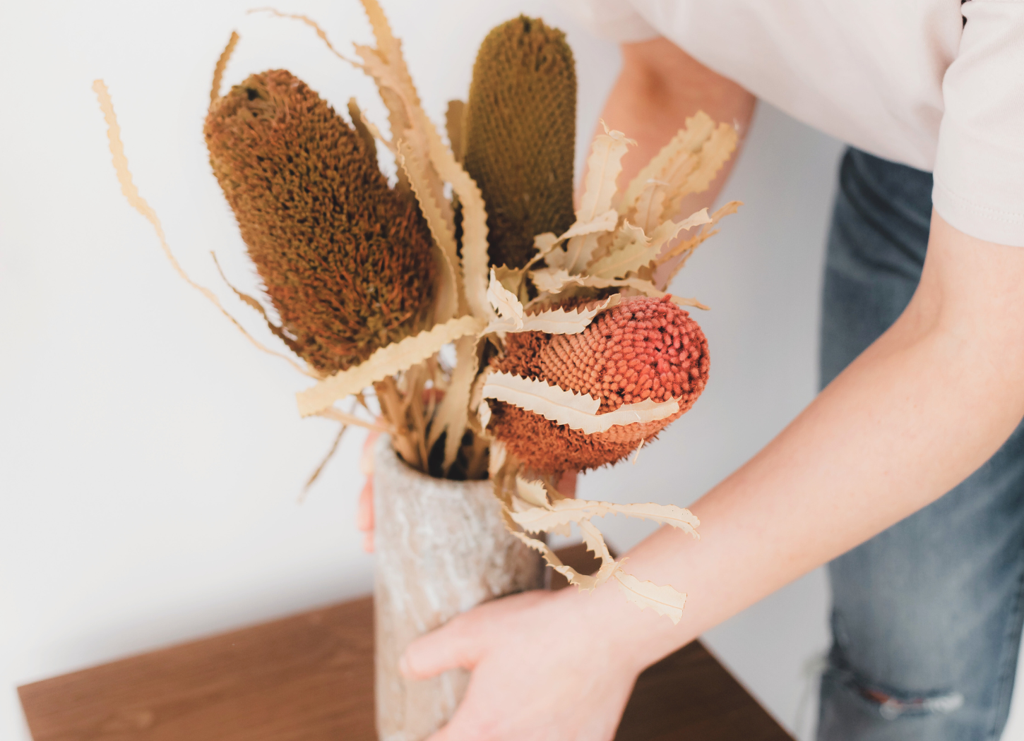 中央に植物が密集して生けられた花瓶を持つ女性の手が写る。アップ構図で、植物の豊かさと手の動きが際立つ。