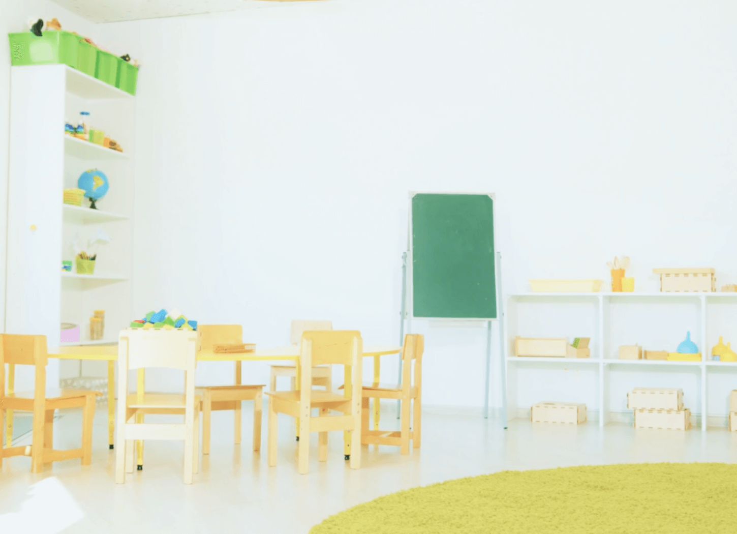 幼稚園の一室。白い壁に小さな木製の子供用椅子と机、中央に縦長の黒板、左右に棚が並ぶ