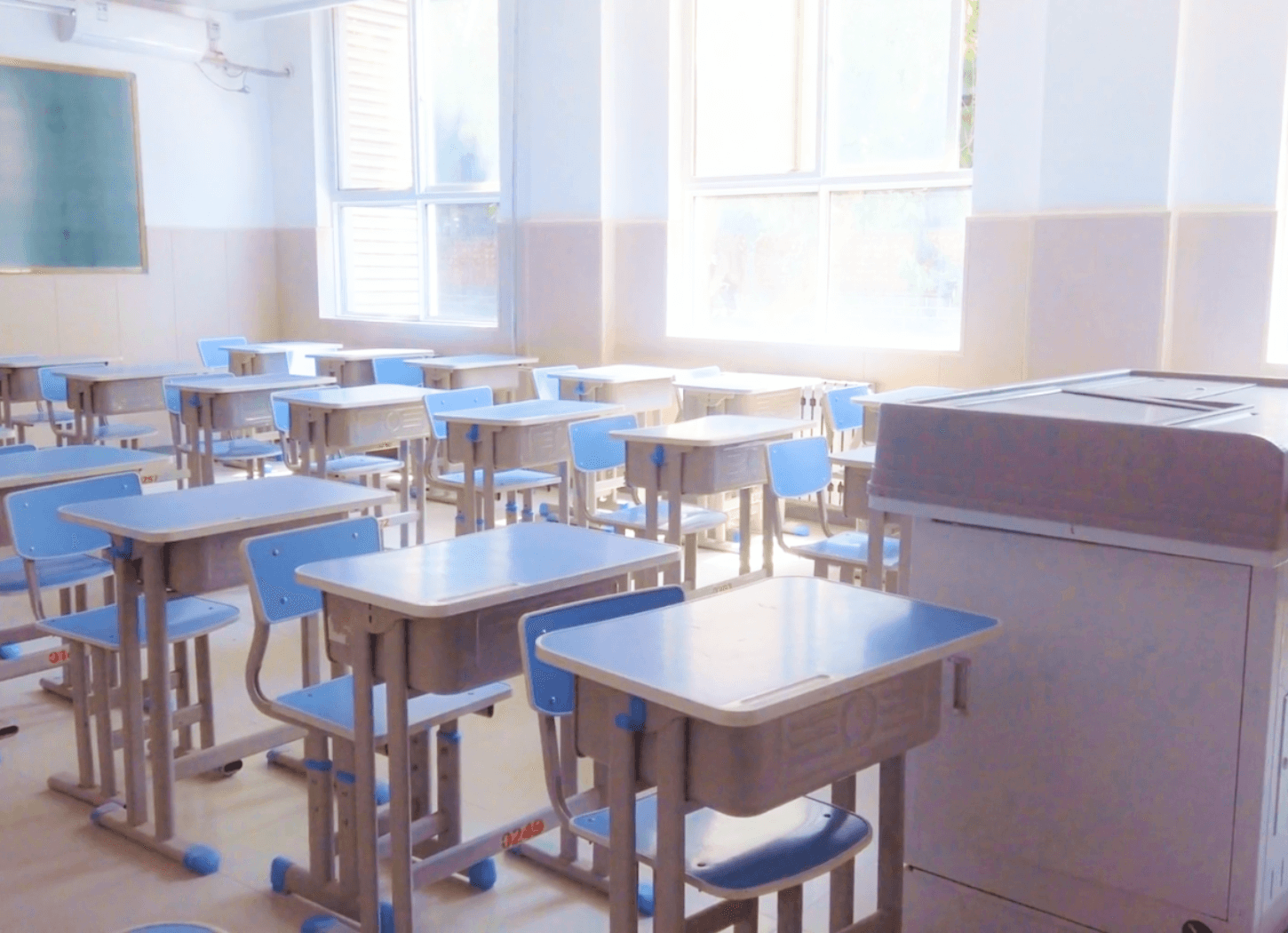 窓から光が差し込む学校の教室。教壇と整然と並ぶ机と椅子が見える