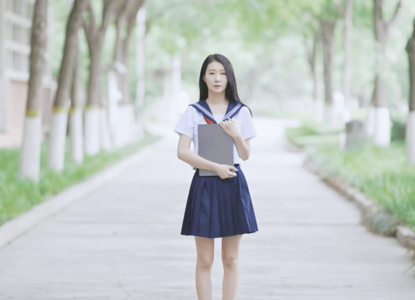 「左右に木が等間隔に並ぶ通りの真ん中で、セーラー服を着た長い黒髪の女の子が本のようなものを胸の前に抱えて立っている