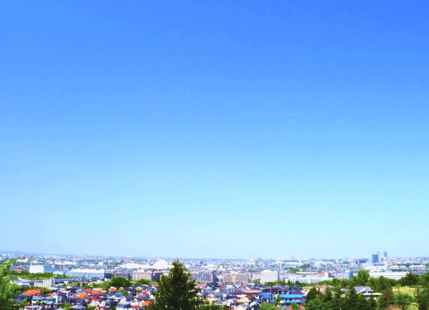 青空が画面の大部分を占め、下方にわずかに木と民家の屋根が見える風景。