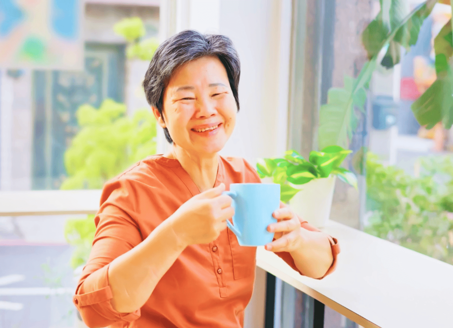 ガラスの壁の前にあるカフェ風のカウンターで、水色の大きなマグカップを持ち、幸せそうに微笑む白髪混じりの女性。おばあちゃんになった「ひろみちゃん」との未来の再会をイメージ。
