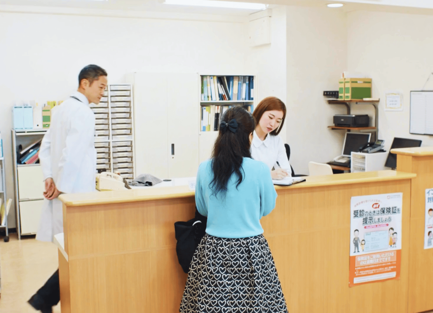 病院の受付で、白衣の女性が向かいに立つ患者の女性に説明をしている様子。その後ろを白衣の男性が通り過ぎる。大人になった二人の「ひろみちゃん」が産婦人科で再会するシーンをイメージ。