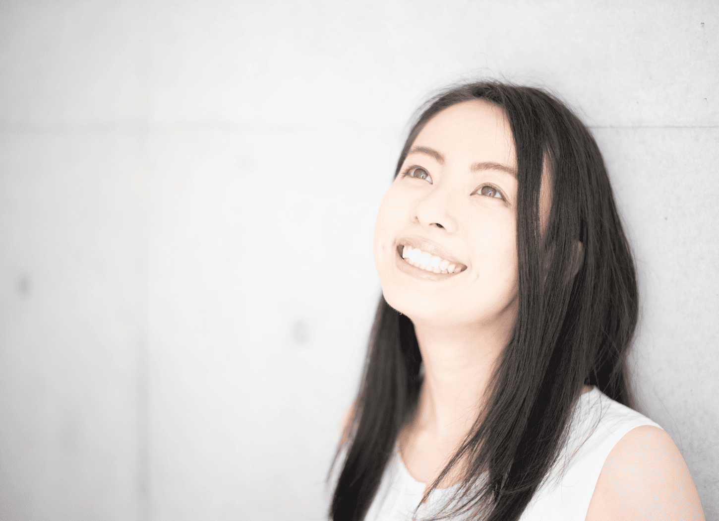 コンクリート打ちっぱなしの壁にもたれて微笑む白いノースリーブの女性。動画撮影や編集の苦手意識を乗り越え、自分なりのスタイルを見つけた瞬間を表現。