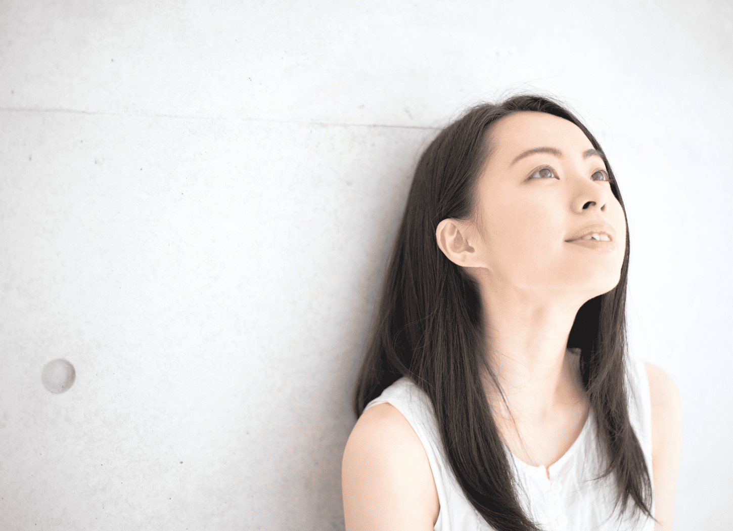 コンクリート打ちっぱなしの壁にもたれる白いノースリーブの女性。長い黒髪で、上空を見つめている。