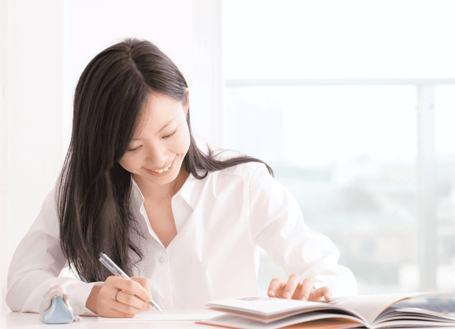 机の上にノートを広げ、笑顔で何かを書き込む白いシャツの女性。背景の大きな窓には遠くの街並みが見える。