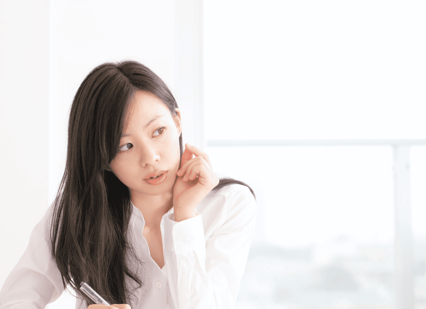 白いシャツを着た黒く長い髪の女性。ノートにペンで書き込みながら、反対の手で頬杖をつき、考え込んでいる様子。背後の大きな窓から遠くの街並みが見える。