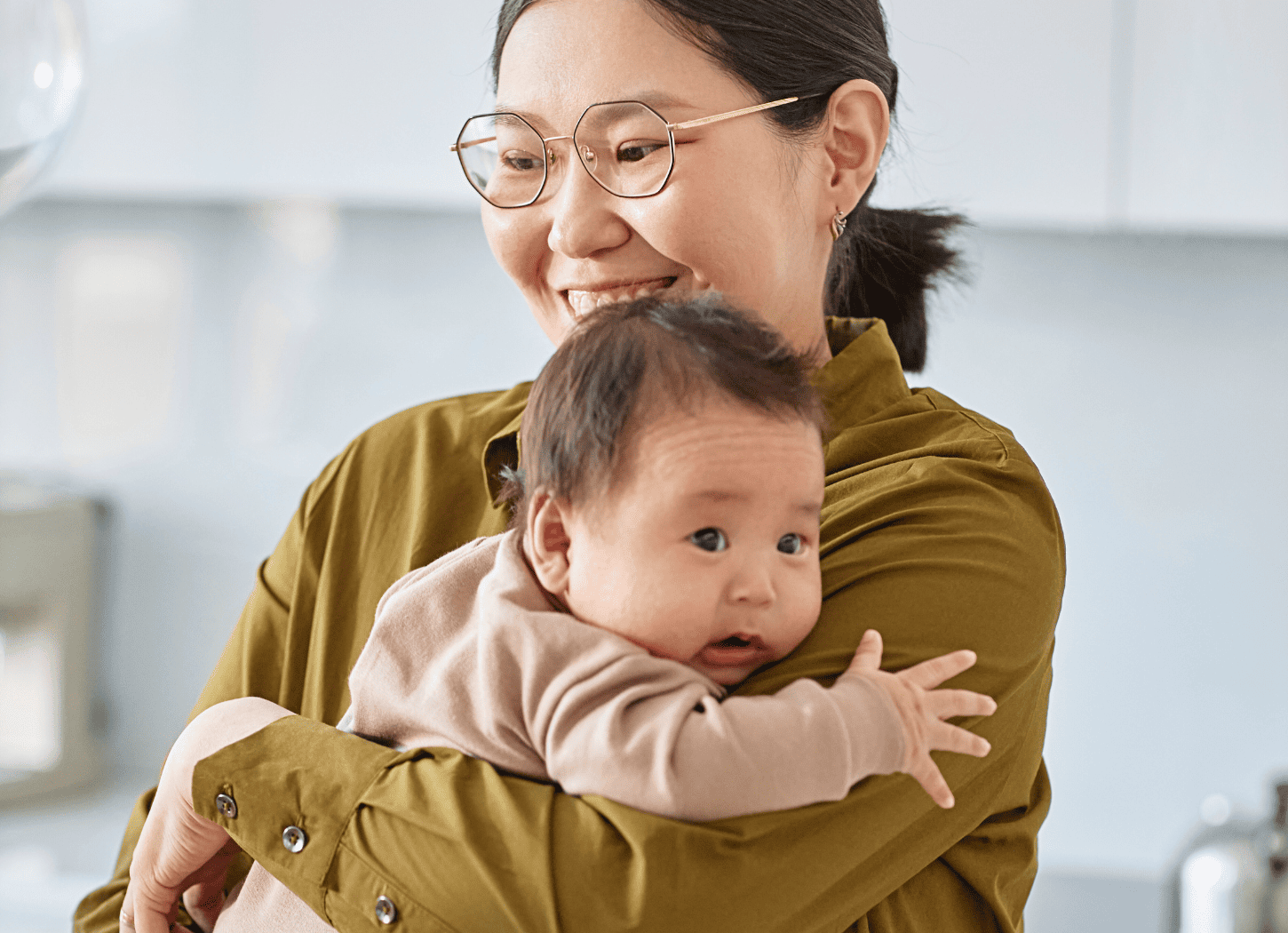 白いキッチンカウンターの前で赤ちゃんを抱っこする女性。シャツを着て黒髪を後ろで一つに結んだメガネの女性と、ベビー服を着た赤ちゃんがこちらを見つめる。育児中のママの日常をイメージ。