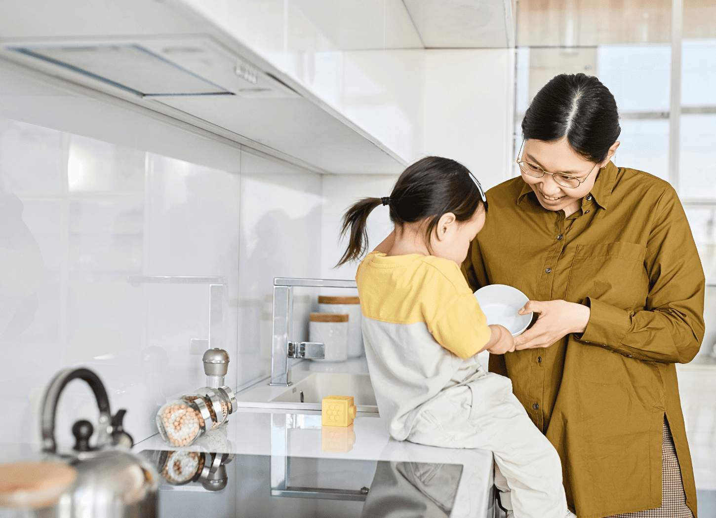 やかんや陶器の容器が見える白いキッチンで、娘を座らせて器を見せる女性。シャツを着て黒髪を後ろで一つに結んだメガネの女性と、好奇心を向ける娘の様子。育児中の親子の日常をイメージ。