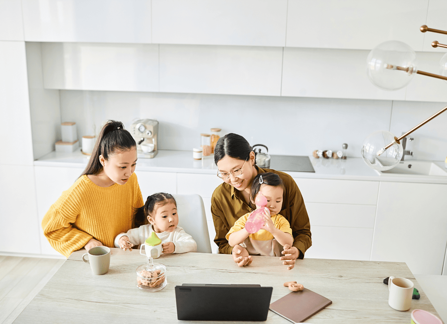 白いキッチン前の大きなダイニングテーブルに、ママ友と並んで座り、お互い膝に娘を乗せてあやしながら話す女性たち。テーブルの上には子供用のマグやお菓子、ノートパソコンなどが置かれている。育児と交流の日常を引きで撮影。