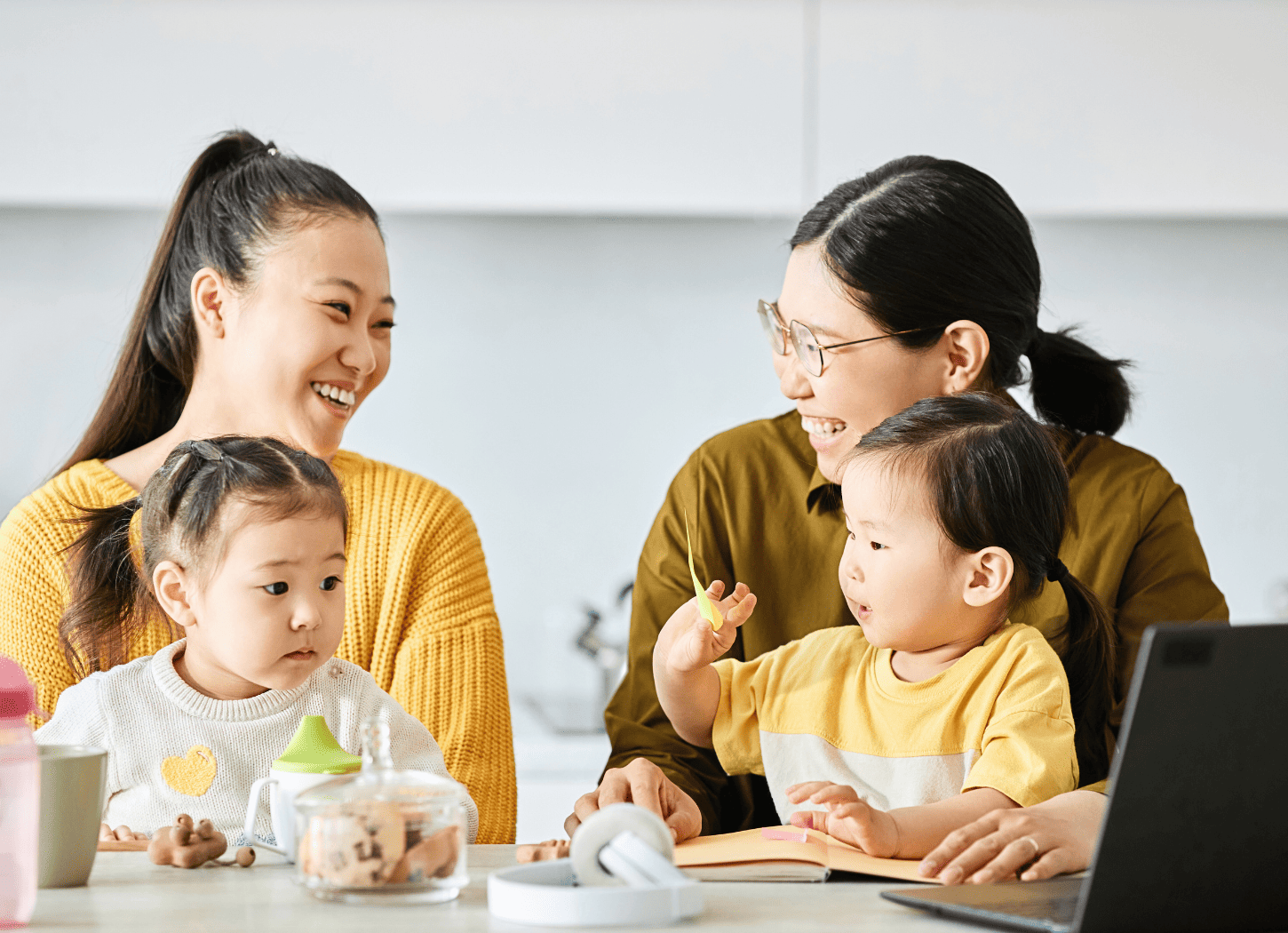 ママ友を自宅に招き、お互いに娘を膝に乗せて談笑する女性たち。白いキッチンカウンターが背景に見える、育児中の交流シーン。