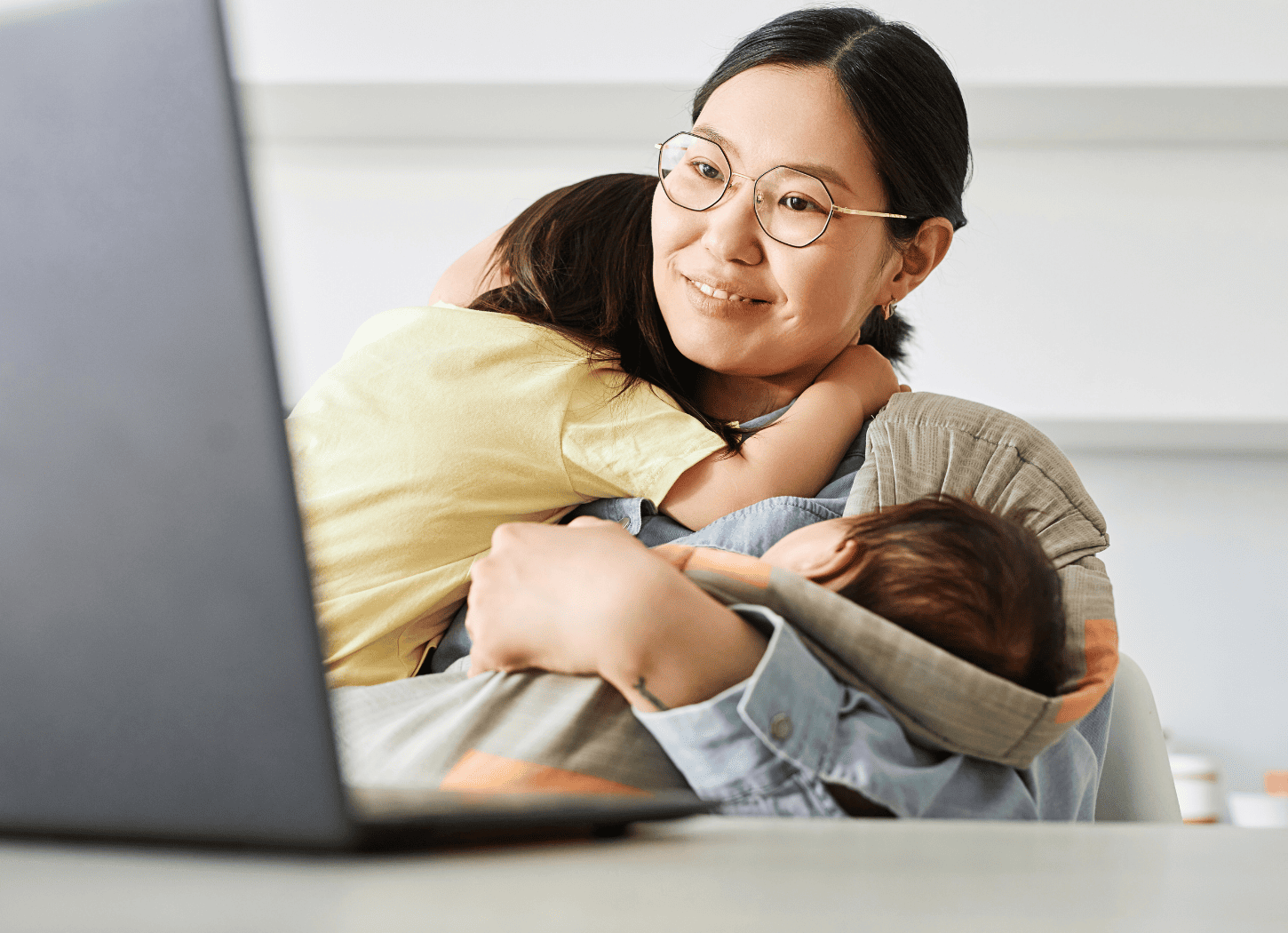 白いキッチンカウンター前のテーブルで、ノートパソコンを広げ、寝ている娘と赤ちゃんを膝に乗せながら画面を見つめる女性。シャツを着て黒髪を後ろで一つに結んだメガネの女性。育児と仕事を両立する日常をイメージ。