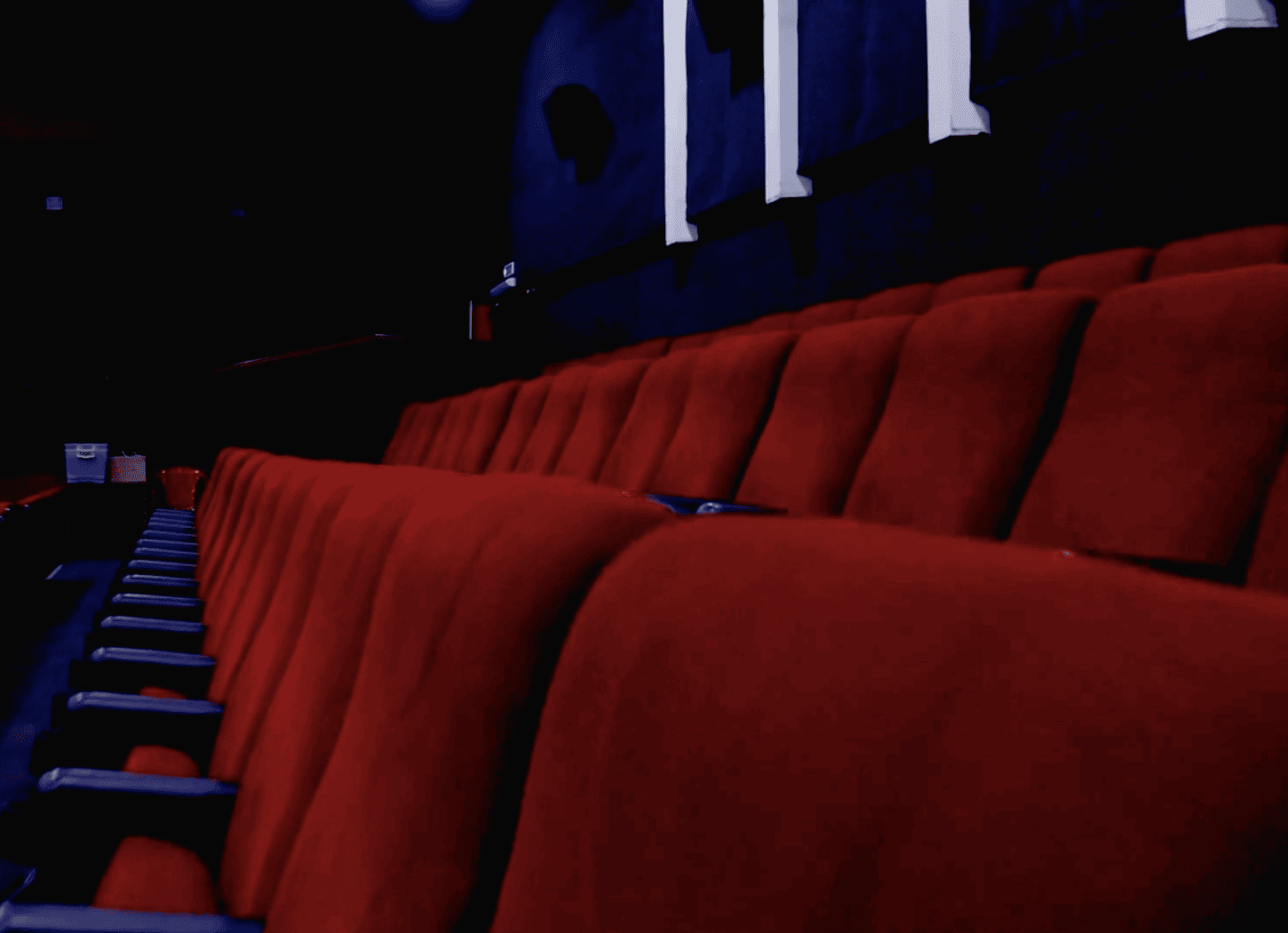 黒い壁と床の映画館で、赤い座席と手すりが横に並ぶ。奥には非常口が見え、暗闇の中で映画館の静けさを感じる情景。