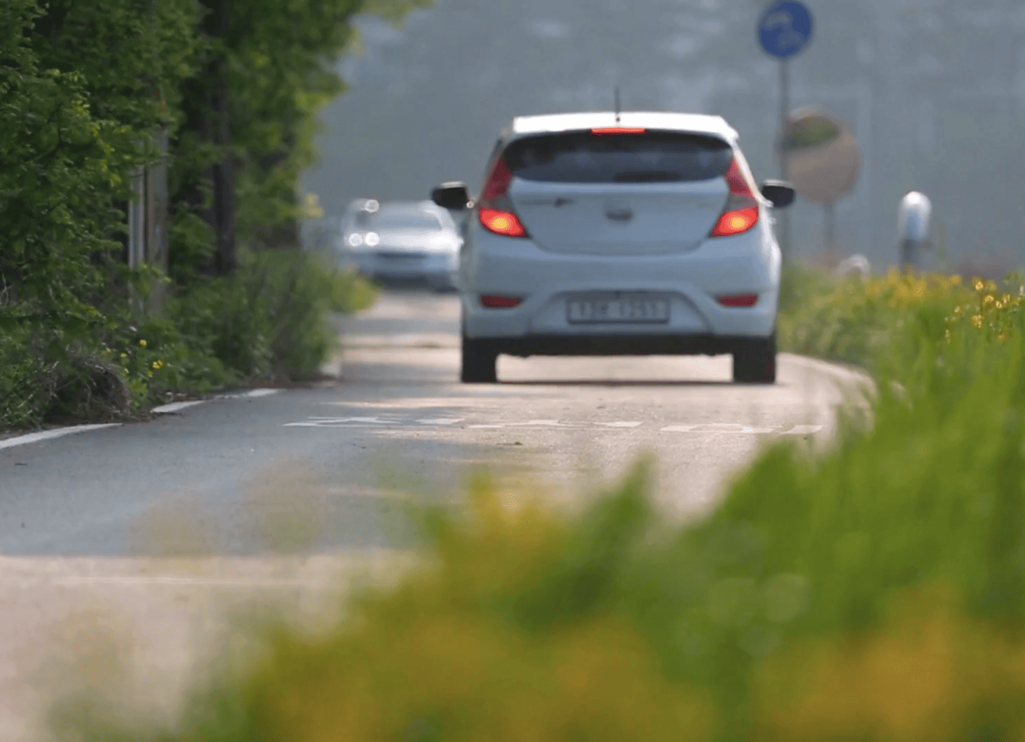 緩やかにカーブするアスファルトの道を走り去ろうとする白い車。左右には草や木が生え、奥には青い標識が見える。Nちゃんの引越しと別れのイメージを象徴する情景。