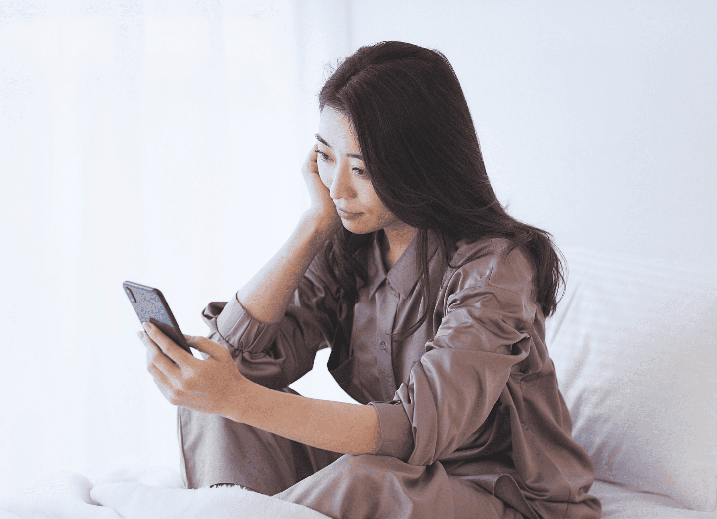 白いベッドの上でパジャマ姿の女性が頬杖をつきながらスマホを見つめる。白いカーテンから柔らかい光が差し込む、何もしない日曜日のリラックスシーン。