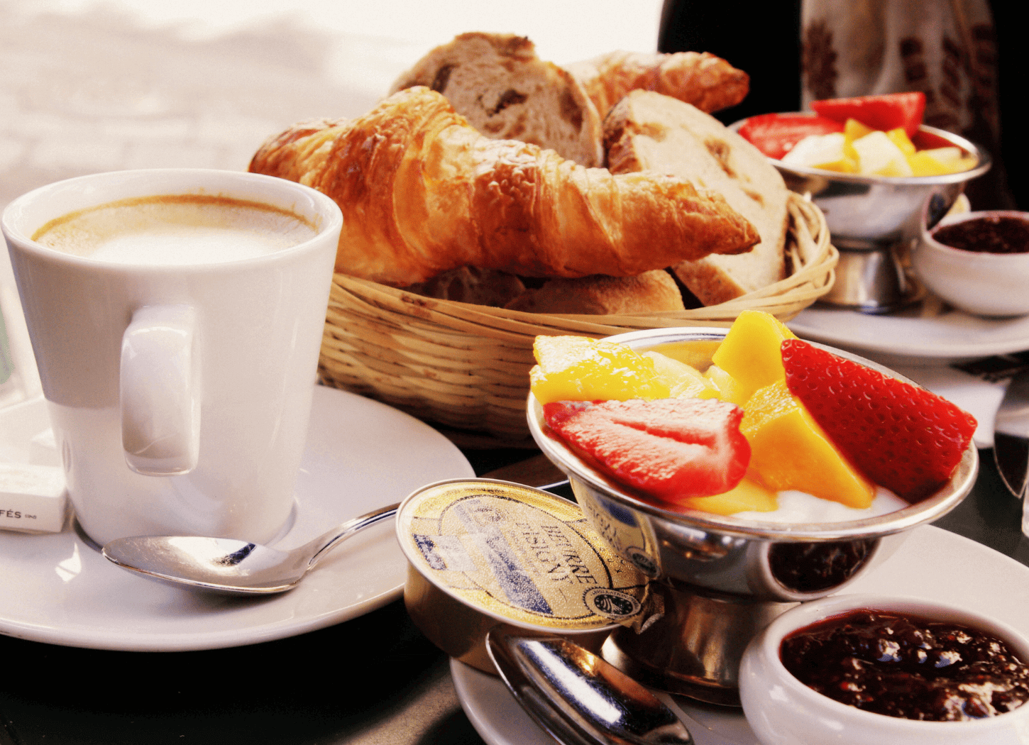 パリのカフェの黒い木製テーブル。カゴに盛られたさまざまな種類のパン、シルバーのボウルに守られた色とりどりのフルーツ、カフェラテの入ったカップが置かれている。