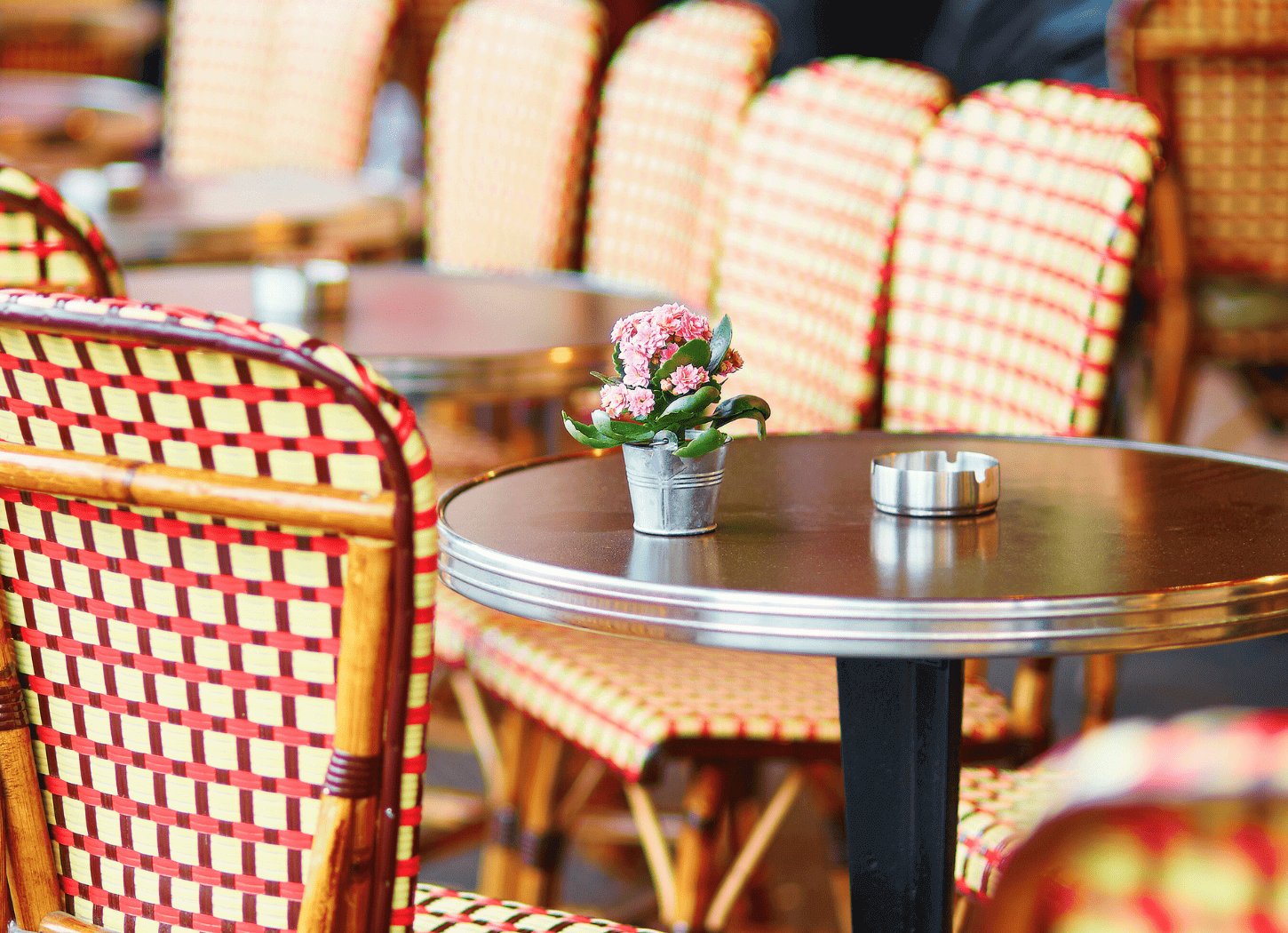 パリのカフェまたはビストロのテラス席。格子柄の椅子と小さなシルバーの円卓が置かれ、卓上にはピンクの花が飾られている。