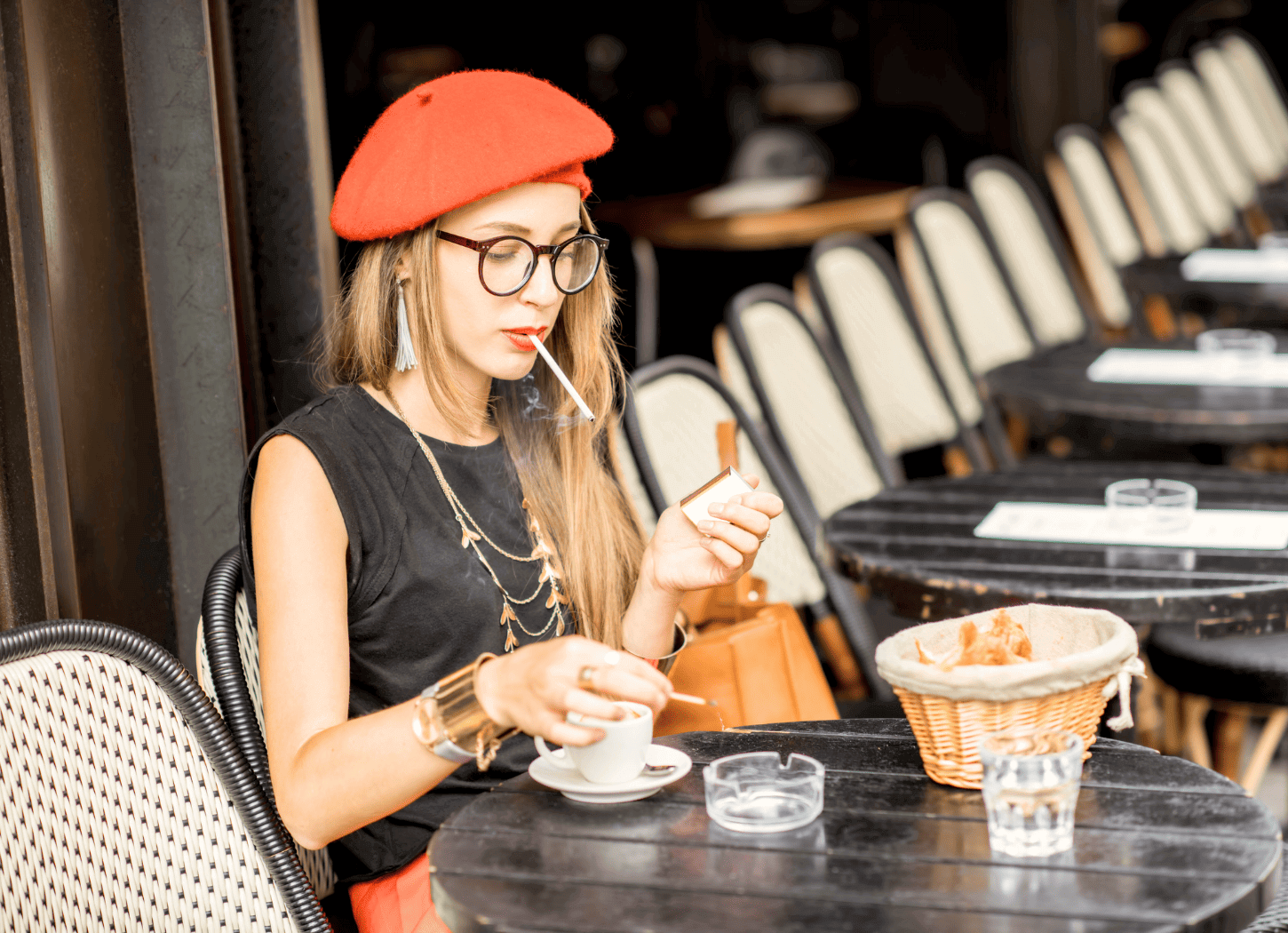 黒で統一されたカフェのテラス席で、タバコを咥えながらコーヒーカップを手に取ろうとしている女性。黒のノースリーブに金髪、赤いベレー帽、黒縁メガネを身につけている。