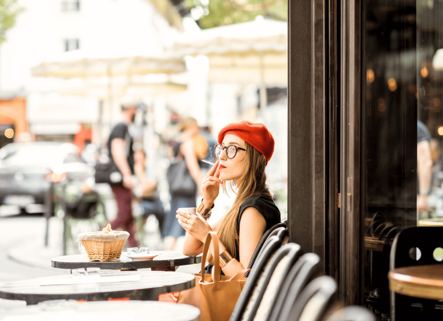 黒で統一されたカフェのテラス席でタバコを吸う女性。黒のノースリーブに金髪、赤いベレー帽、黒縁メガネを身につけ、遠くを眺めている。