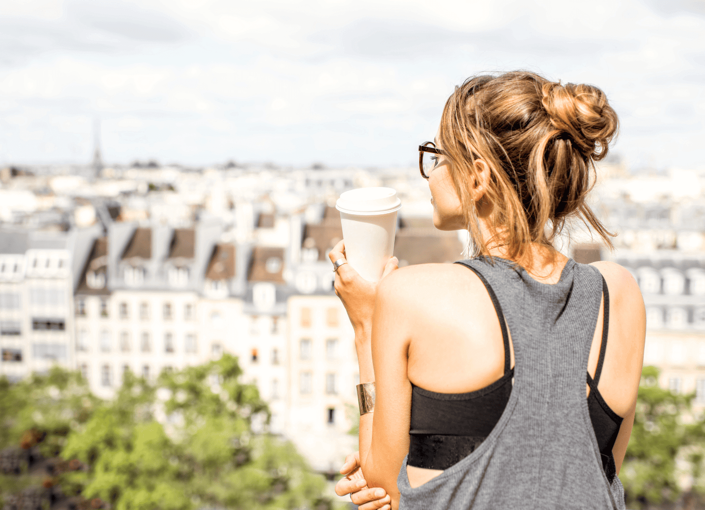 金髪をお団子にした女性の後ろ姿。黒縁の眼鏡をかけ、ノースリーブの服を着て、紙のタンブラーを手にパリの街並みを眺めている。
