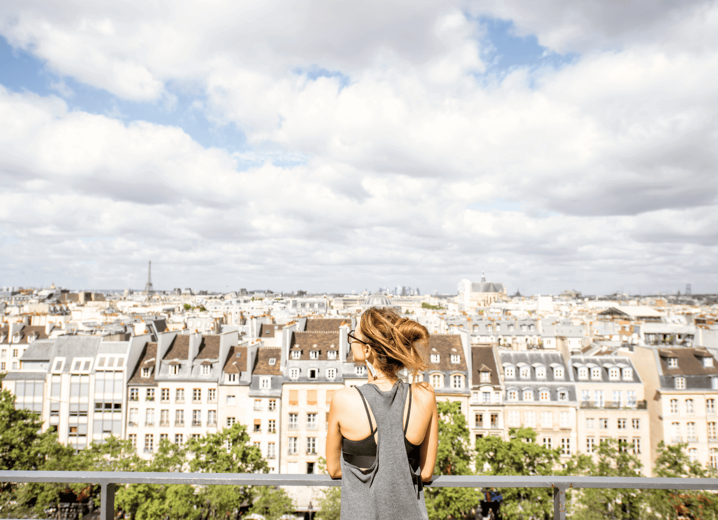 金髪をお団子にした女性の後ろ姿を引きで撮影。手には紙のタンブラー、目の前には柵があり、青い空に浮かぶ雲の下にパリの街並みが広がっている。