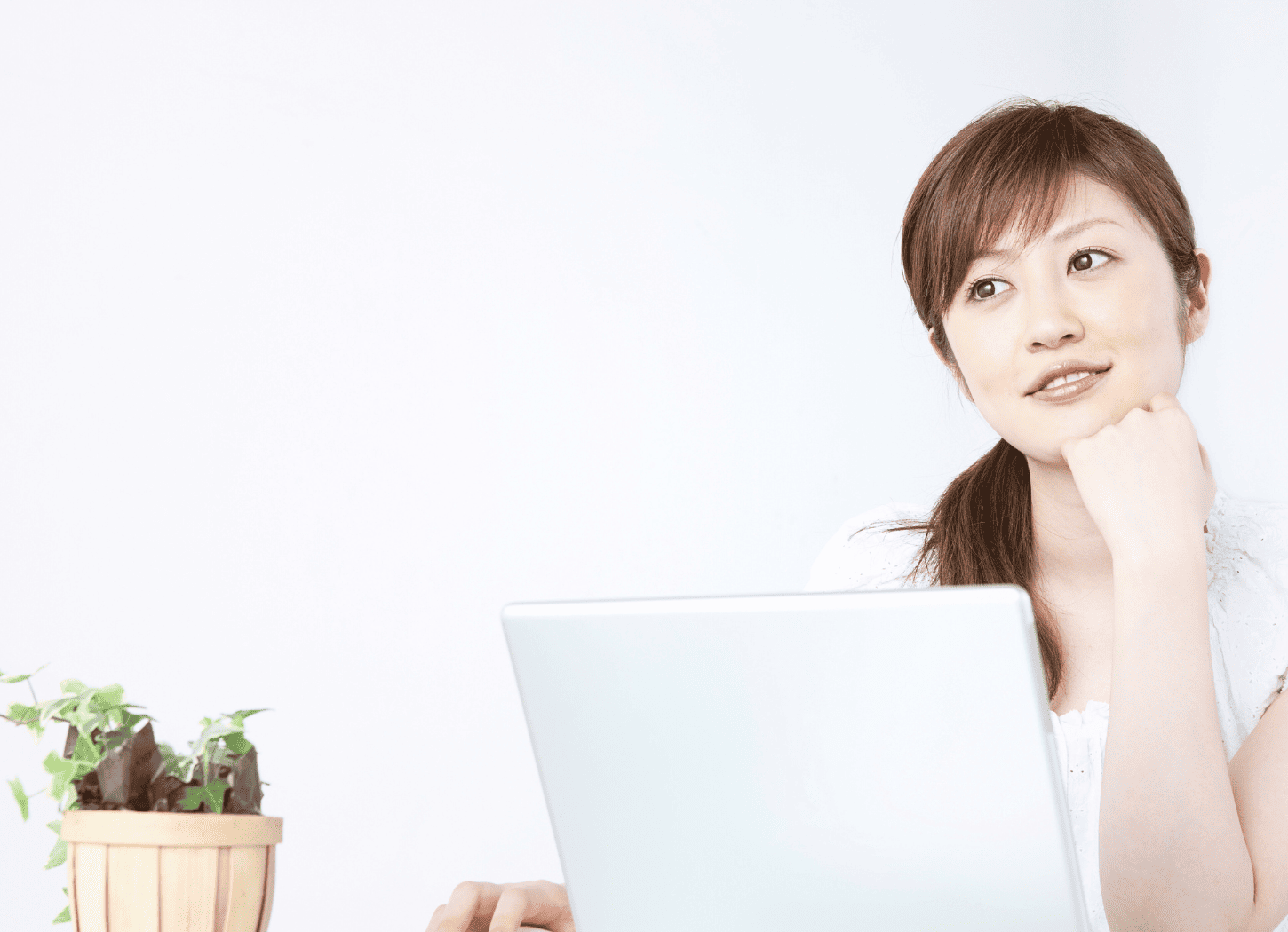 白い服を着た女性が白いテーブルにノートパソコンを開き、ナチュラルな鉢植えの小さな観葉植物が置かれている。女性は頬杖をついて横を見ており、正面から撮影されている
