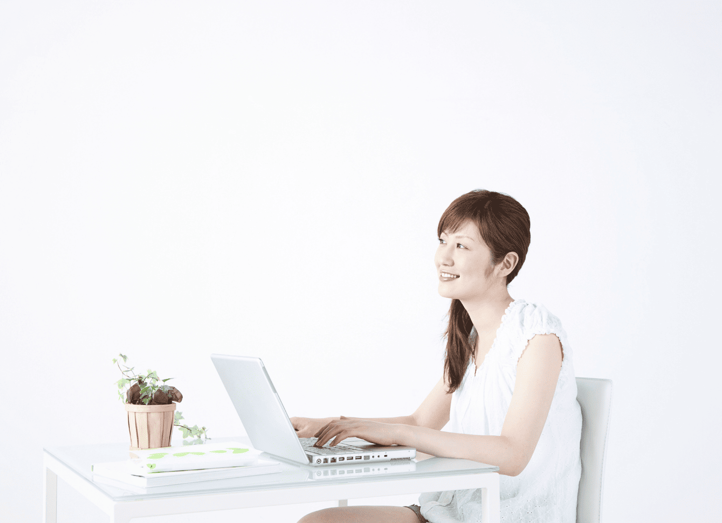 白い服を着た女性が白い壁の前で、ガラス製のテーブルに置かれたノートパソコンを前に斜め前を見つめて話しかけている。両手はキーボードの上にあり、テーブルには観葉植物と資料が置かれている。横から引きで撮影されたシーン
