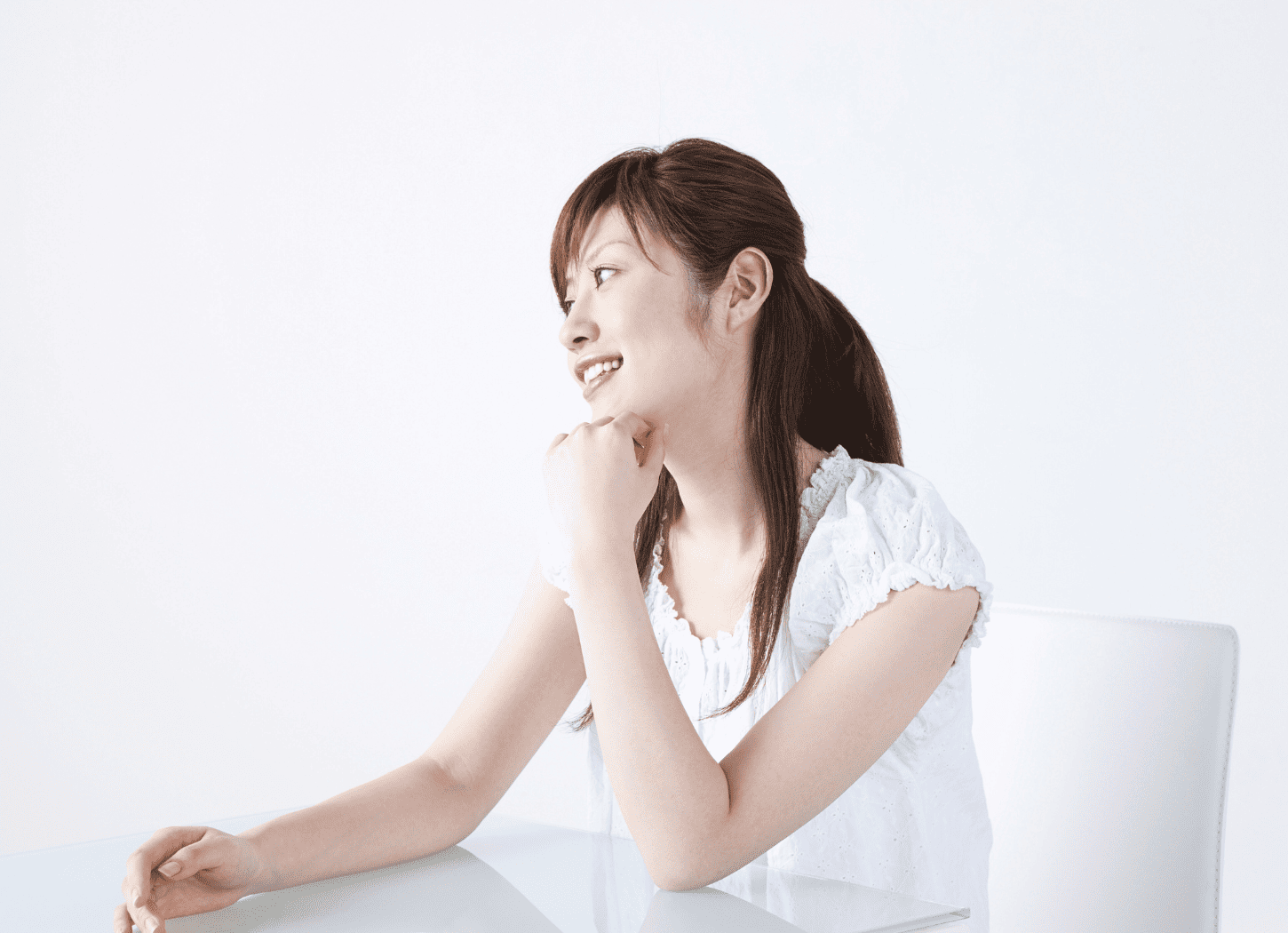 白い服を着た女性が白い壁の前に置かれたガラステーブルに頬杖をつき、横を向いて話している様子。白い椅子に座っている
