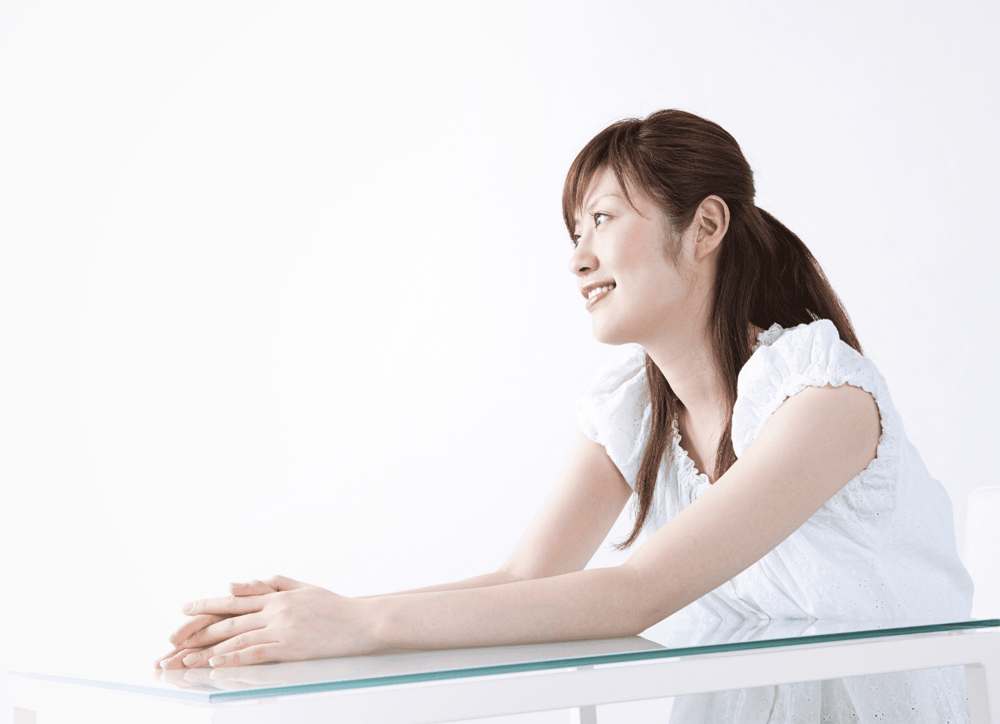 白い服を着た女性が白い壁の前のガラス製テーブルに座り、白い椅子に腰掛けて誰かに向かって話している様子。斜め下からのアングルで撮影されている
