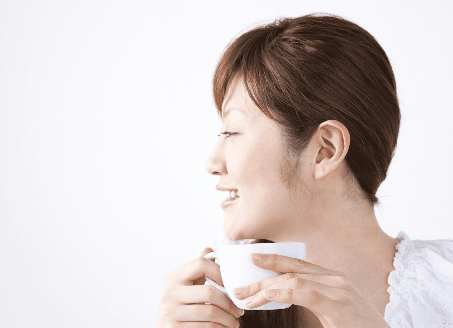 白い服を着た女性の横顔のアップ。白い壁の前で白いティーカップを両手で持ち、飲み物を口にしながら笑顔で談笑している様子
