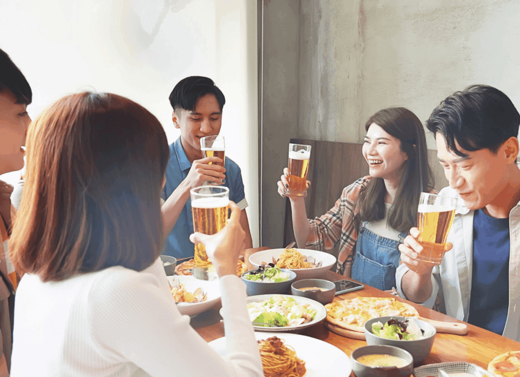 グラスを傾けてビールを飲む男女6人。テーブルにはパスタやサラダが並び、昼のレストランで笑顔が広がっている。