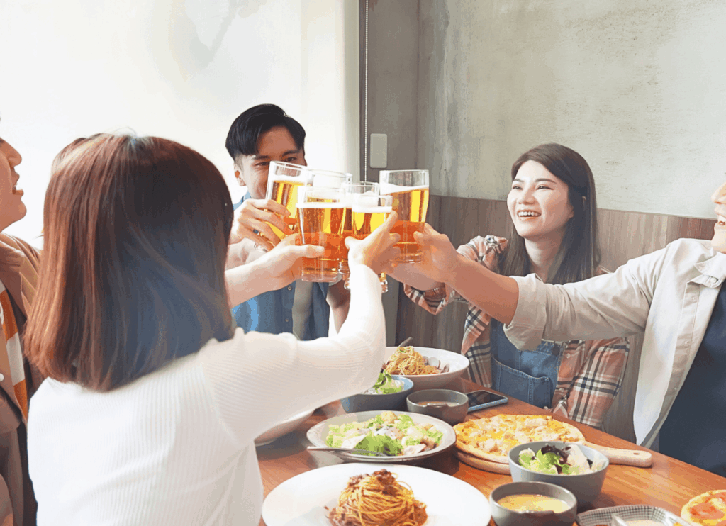 仲良しの男女6人がグラスを片手に乾杯している。テーブルにはパスタやサラダが並び、明るい昼のレストランで笑顔があふれている。