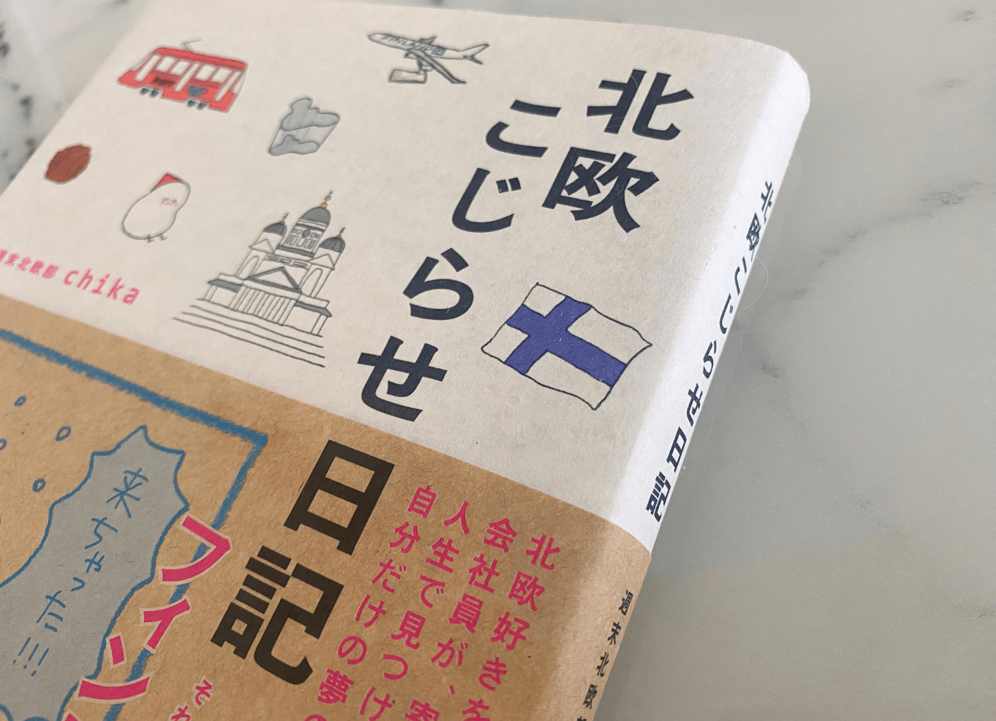 週末北欧部 chikaのコミックエッセイ『北欧こじらせ日記』の表紙を斜めから撮影した写真。タイトルの文字が大きくはっきり見え、北欧らしいイラストが優しい雰囲気で描かれている。
