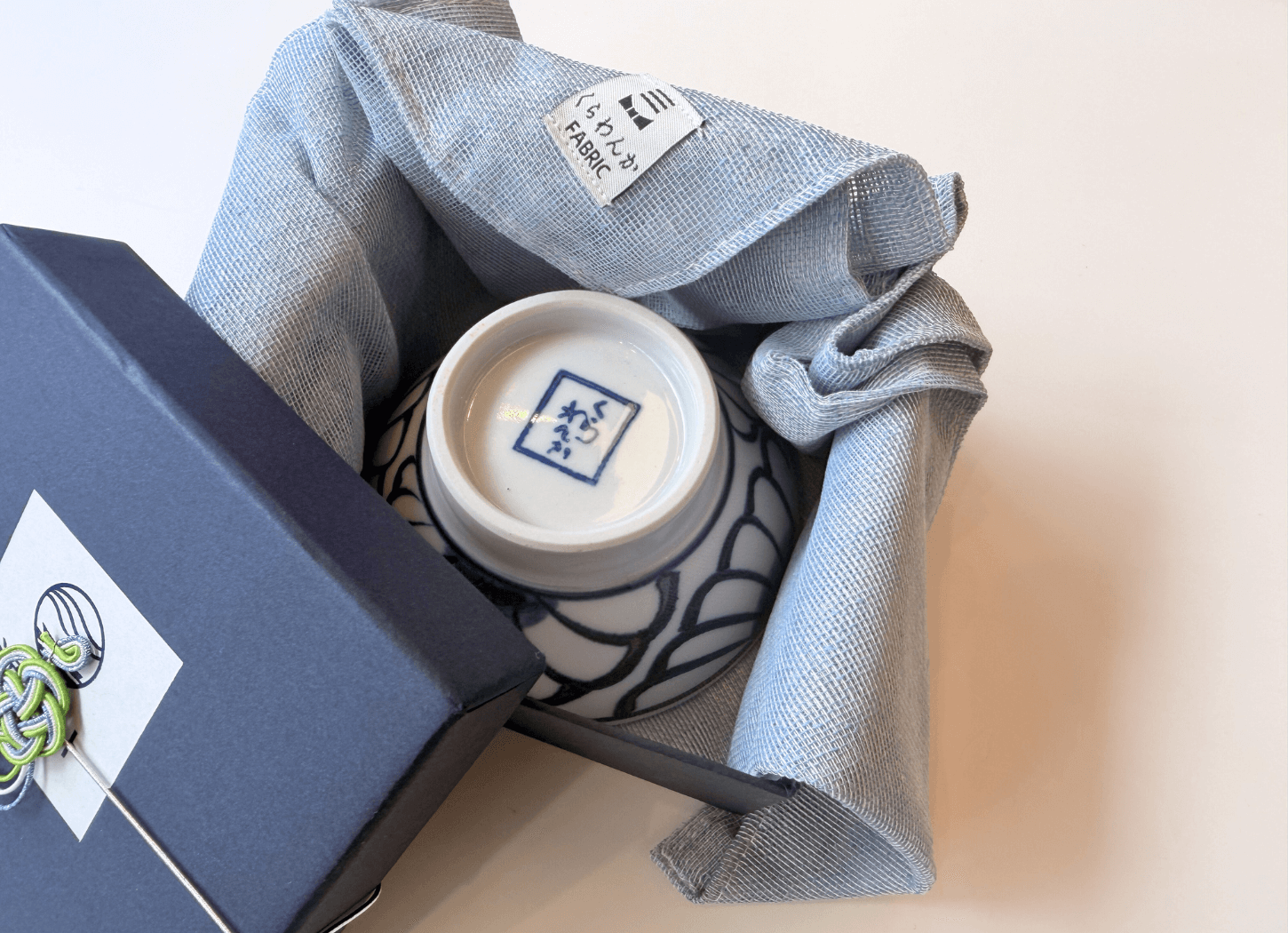 波佐見焼の白磁の器と、くらわんかファブリックのヨシおてふきんを組み合わせた復刻コラボセット。紺色のギフトボックスに収められた様子