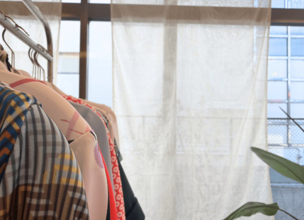 くらわんかファブリックのヨシ素材カーテン(ホワイト)が窓辺に吊るされたde lusso店内。手前には色鮮やかな洋服が並ぶ様子