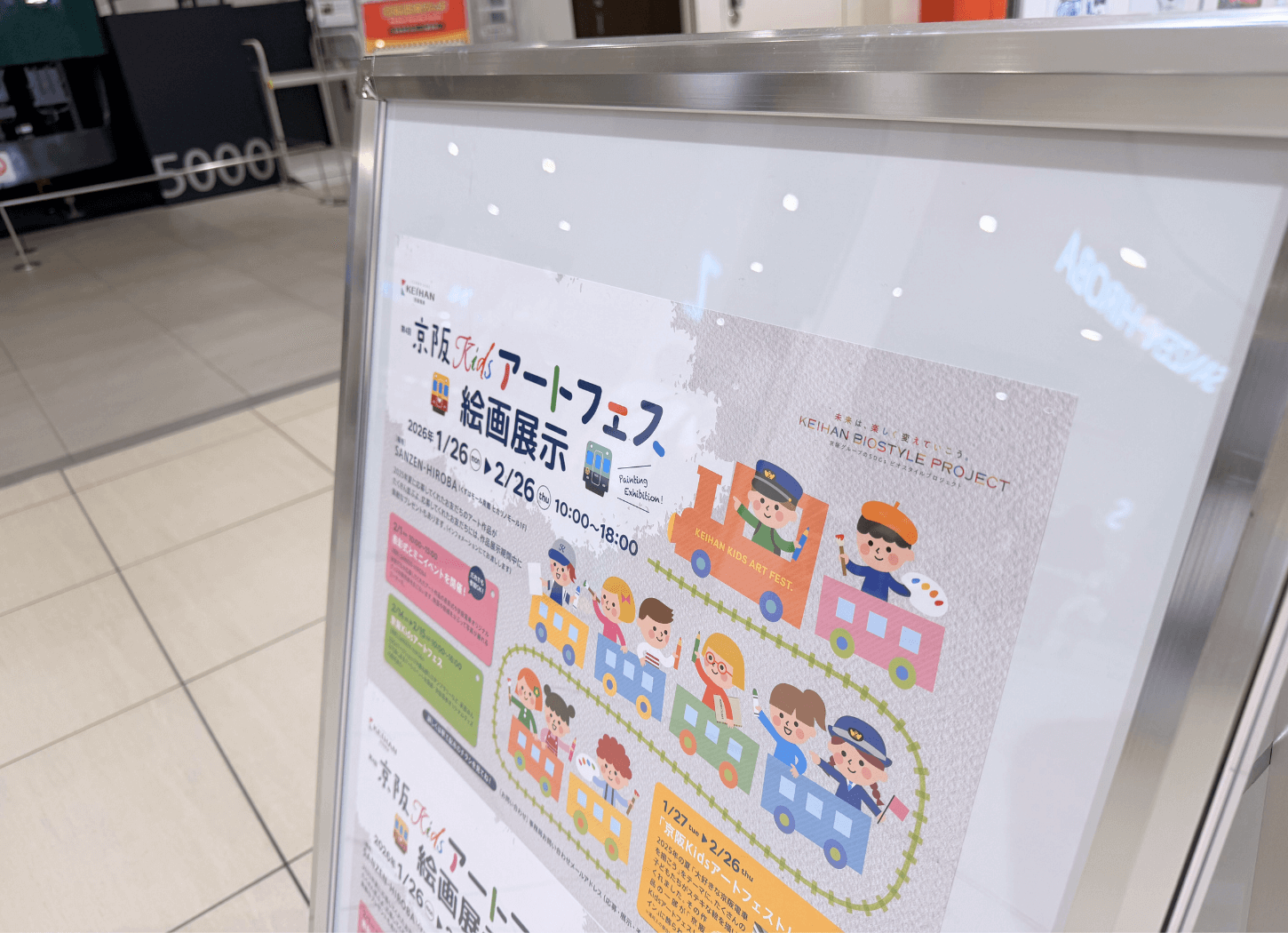 くずはモールSANZEN-HIROBAに掲出された第4回京阪Kidsアートフェスのポスター。制帽をかぶった子どもが列車の先頭に立ち、後ろの車両に絵筆やクレヨンを持った子どもたちが描かれているイラスト。