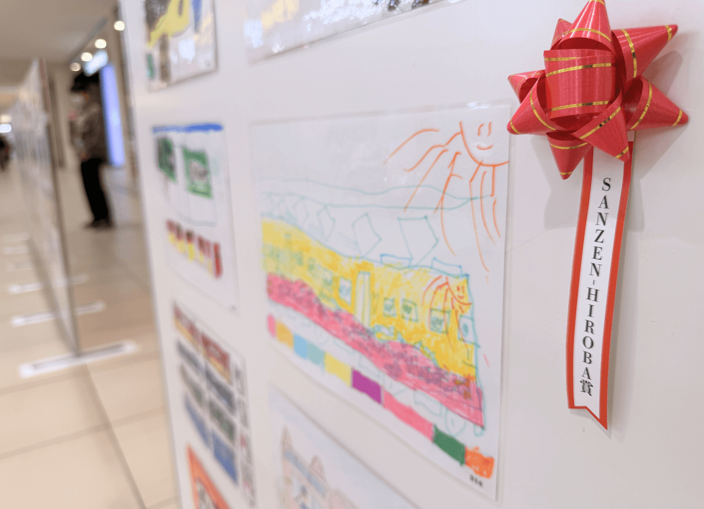 くずはモールSANZEN-HIROBAに並ぶ第4回京阪Kidsアートフェスの受賞作品。白い画用紙に描かれた京阪電車のイラストと、「SANZEN-HIROBA賞」と書かれた赤いリボンが付いた展示の様子。