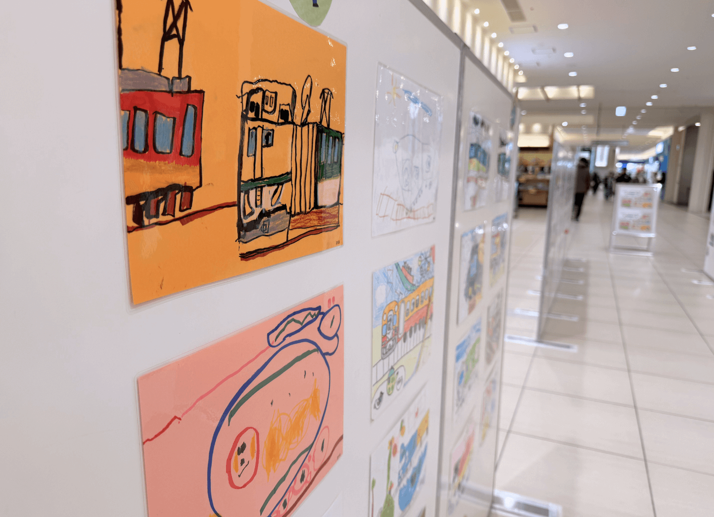 くずはモールSANZEN-HIROBAに並ぶホワイトパネルに、第4回京阪Kidsアートフェスの応募作品が展示されている様子。子どもたちが描いた京阪電車のイラストが並んでいる。