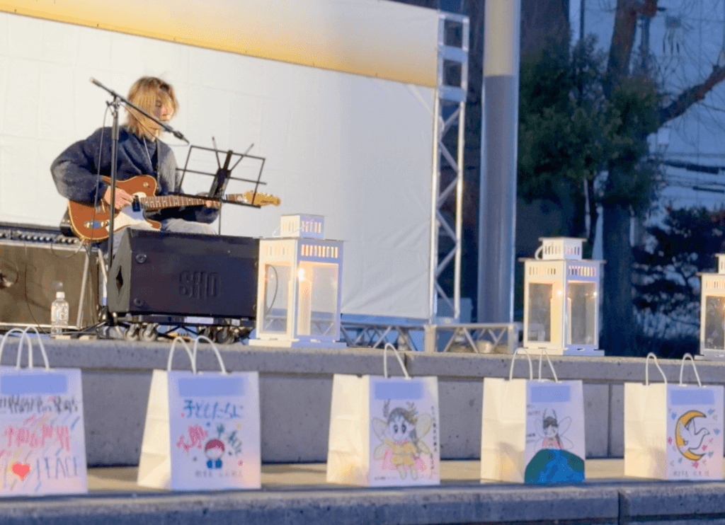 ニッペパーク岡東中央の野外ステージで男性が弾き語りをし、手前にキャンドルが並んでいる様子